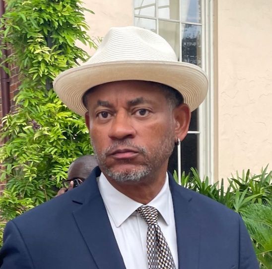 Man in a blue suit and white hat standing outdoors, surrounded by greenery.