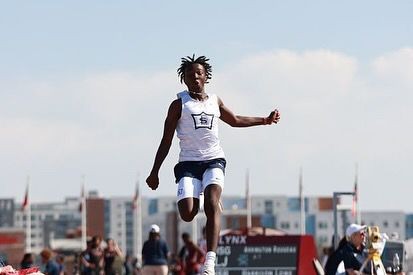 Athlete mid-air during a long jump event with a cityscape and spectators in the background.