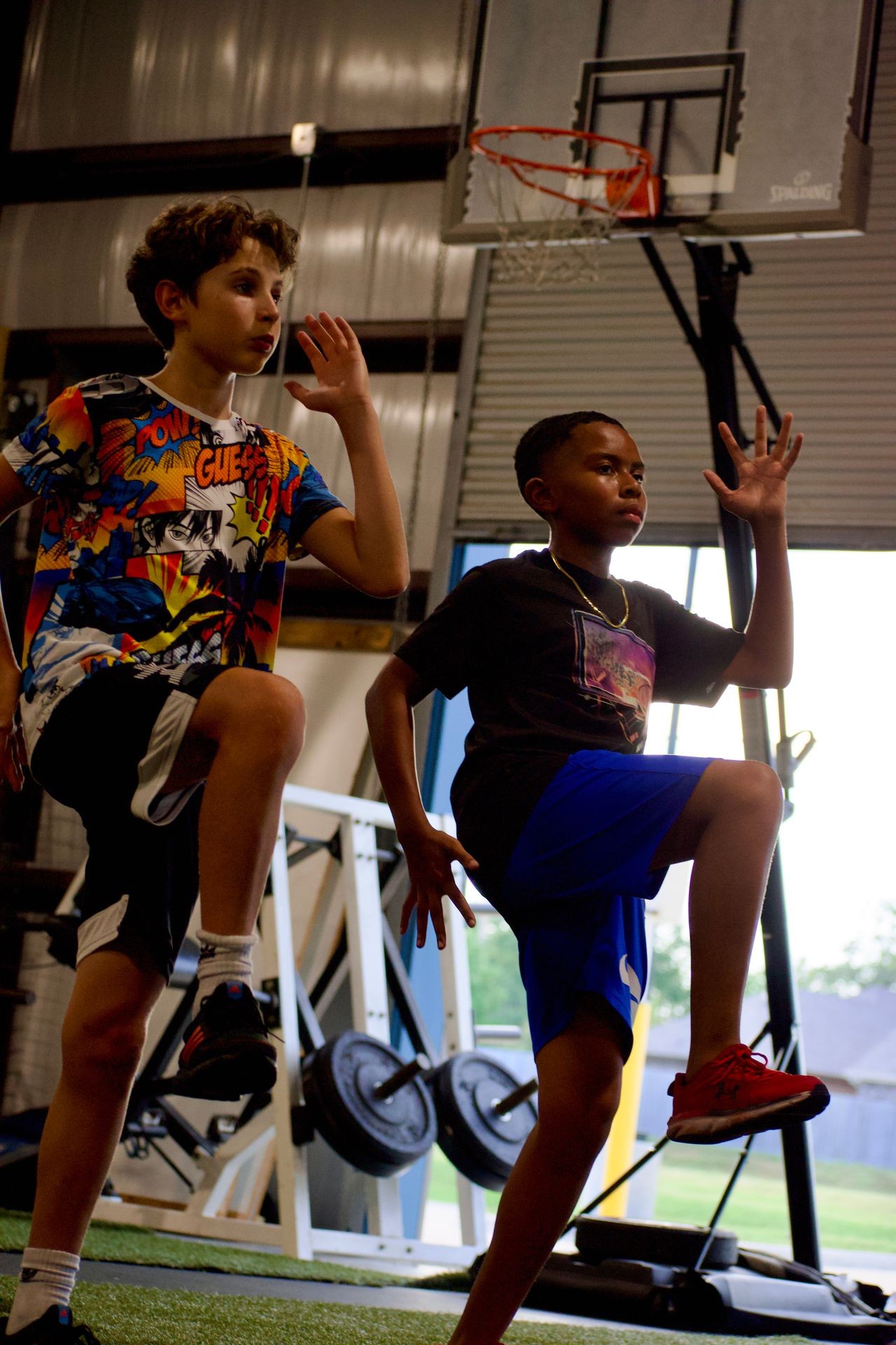 Two boys performing warm-up exercises in a gym with a basketball hoop in the background.