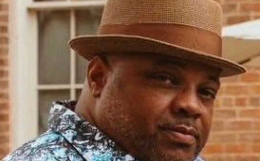 Person wearing a brown hat and a patterned shirt standing in front of a window and brick wall.