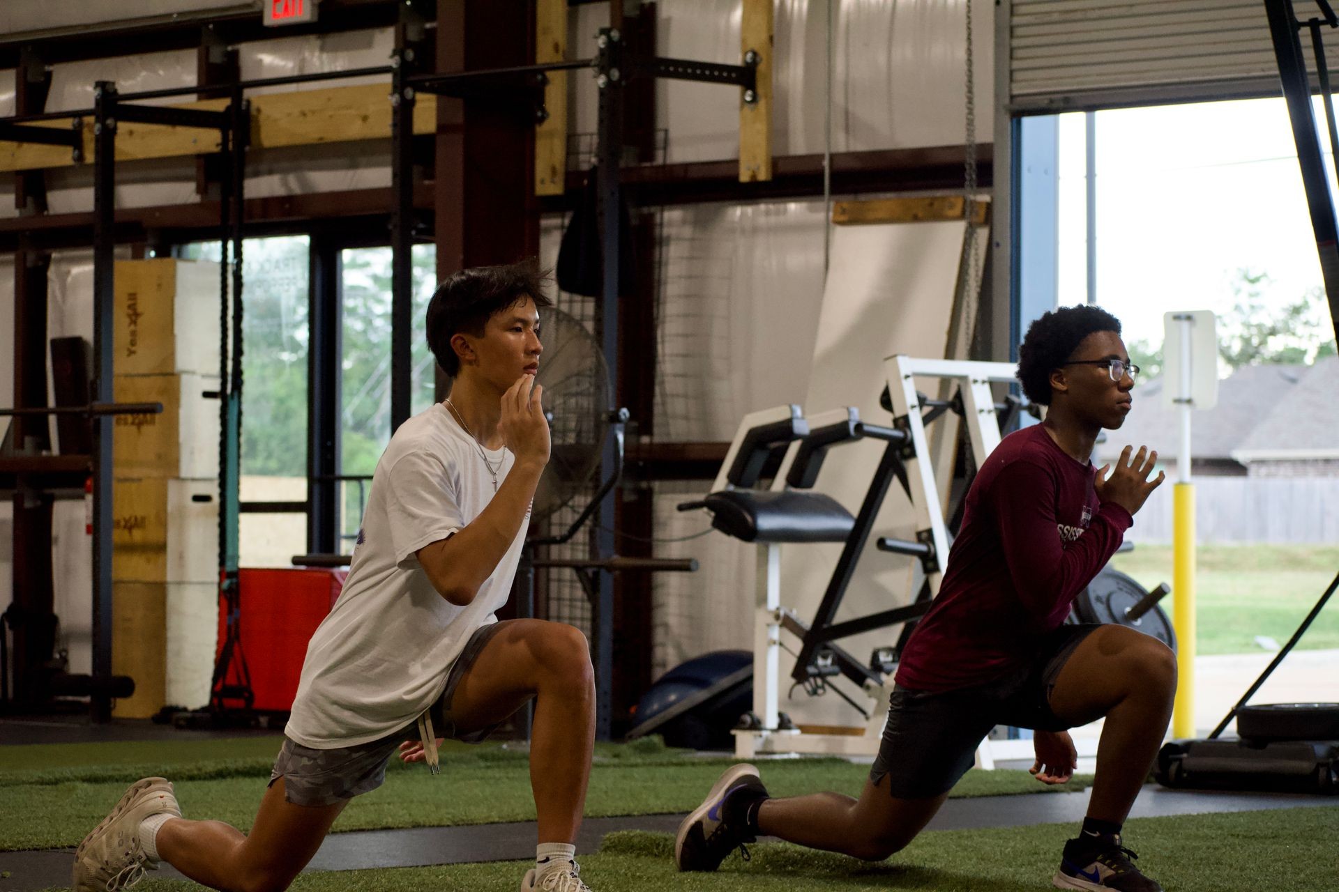 Two individuals performing lunges inside a gym with various exercise equipment visible in the background.