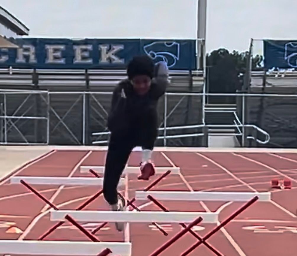 Athlete in motion clearing a hurdle on a track field with a school banner in the background.