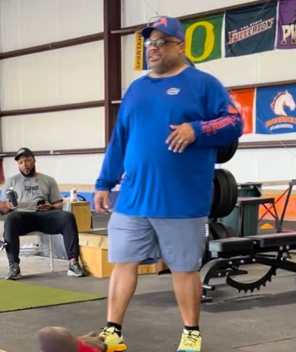 Man in casual sportswear standing in a gym with flags and exercise equipment in the background.